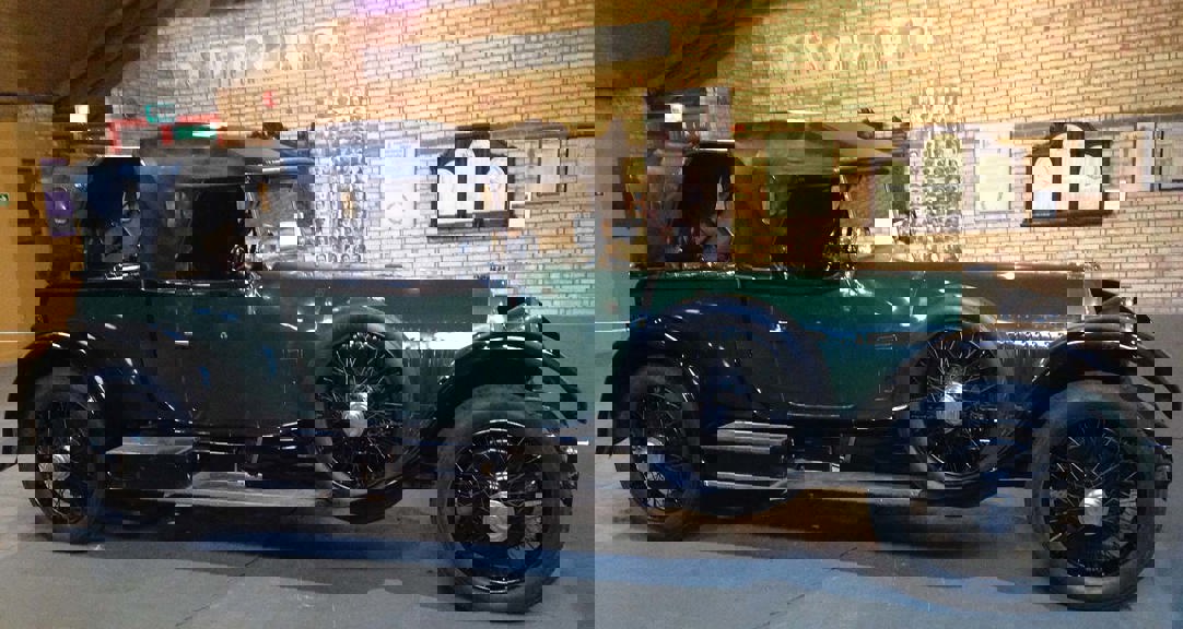 Side elevation of a large vintage car in dark green with a black leather convertible roof, chromed lamps and spoked wheels.