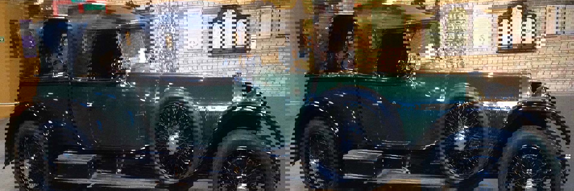 Side elevation of a large vintage car in dark green with a black leather convertible roof, chromed lamps and spoked wheels.
