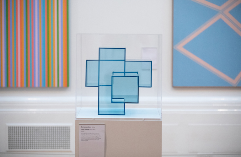 A photograph of an blue geometric sculpture in a display case in an art gallery. In the background there are two brightly coloured abstract artworks,  