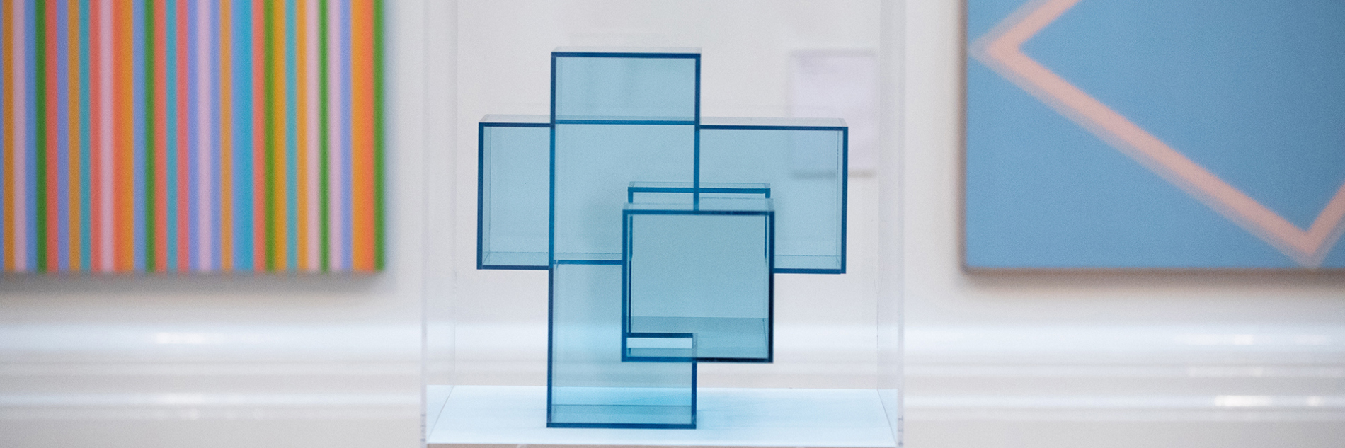 A photograph of an blue geometric sculpture in a display case in an art gallery. In the background there are two brightly coloured abstract artworks,  