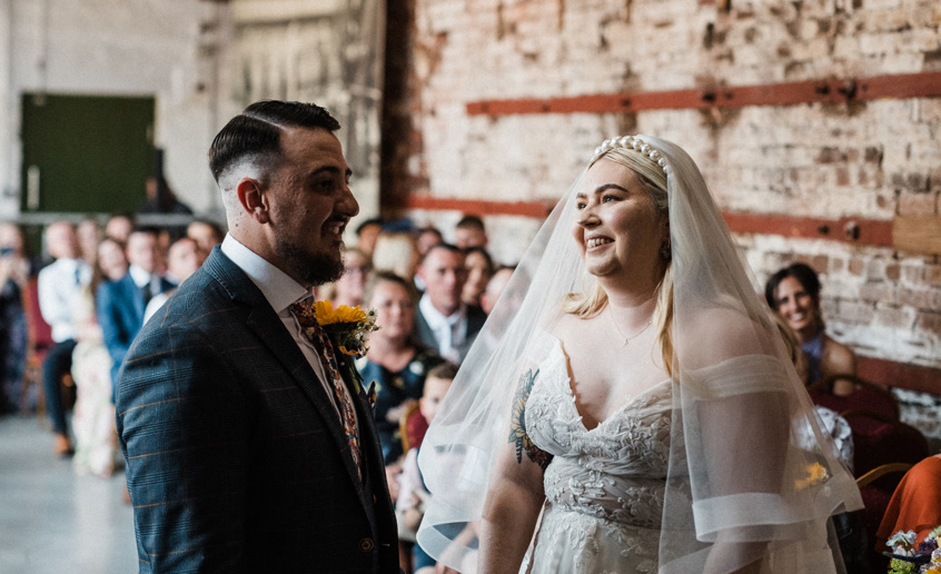 Bride and groom smiling together during wedding ceremony inside an industrial-looking hall.