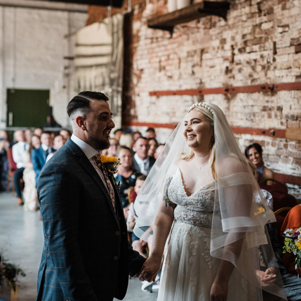 Bride and groom smiling together during wedding ceremony inside an industrial-looking hall.