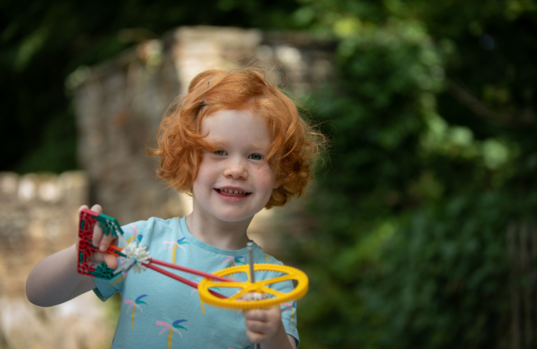 A photograph of a child holding a toy. In the background there are tress and buildings. 