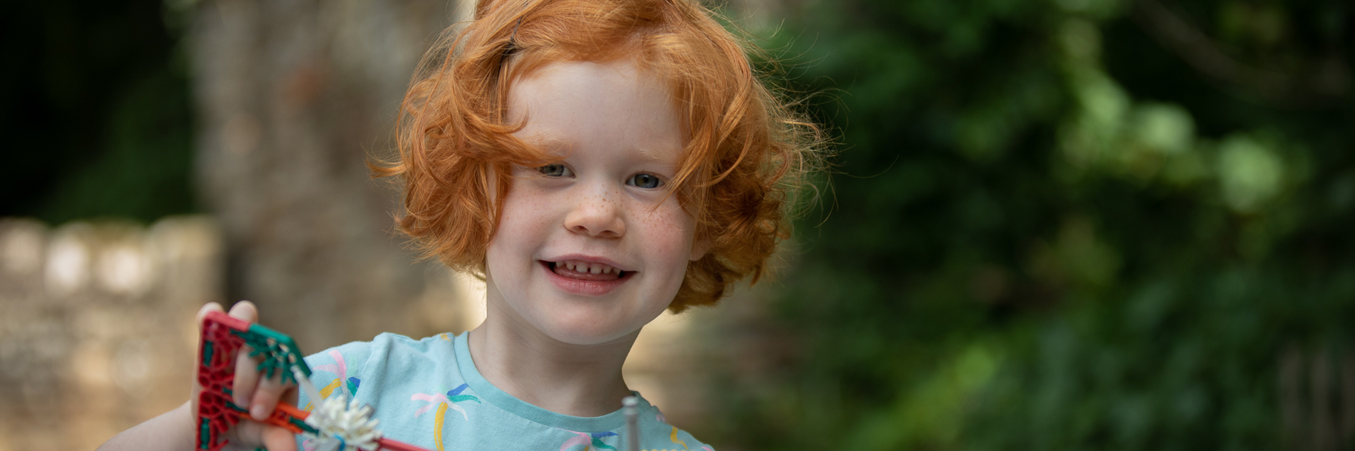 A photograph of a child holding a toy. In the background there are tress and buildings. 