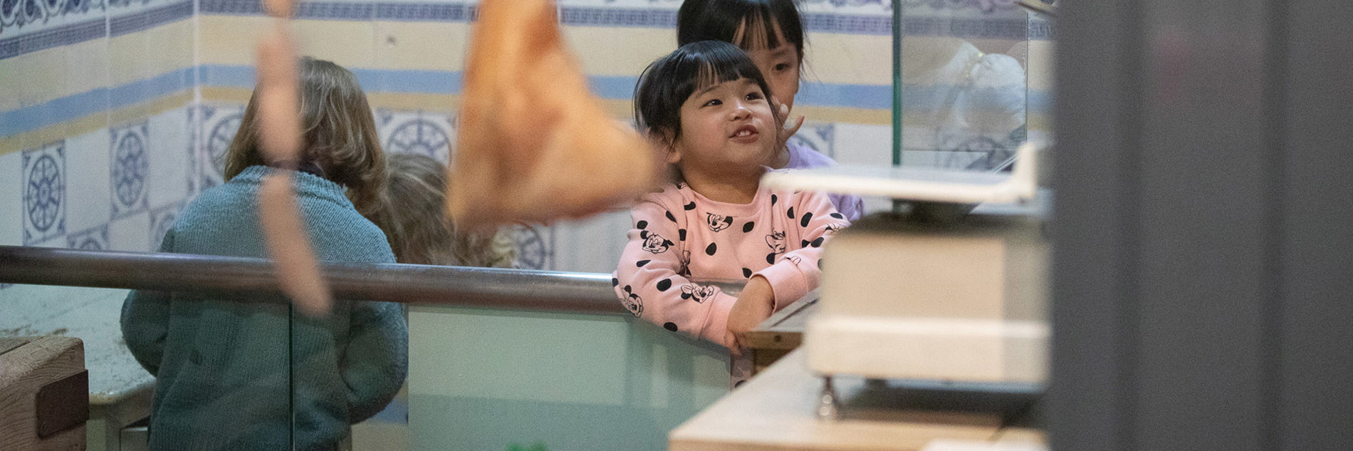 Four young children in the museum's shop recreation, one smiling and leaning against a glass counter. There are plastic meat products in trays in the foreground with sausages and legs of meat hanging down with a background of  blue and white patterned tiles.