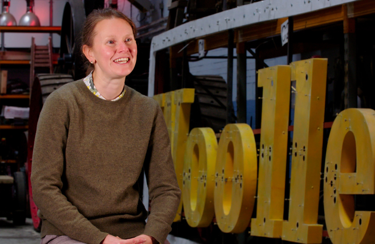 A photograph of a adult sitting in a workshop. There is a large lettered sign next to them (the letters o-l-l-e can be seen) and there are many objects behind them including industrial lights, wheels and ladders. 