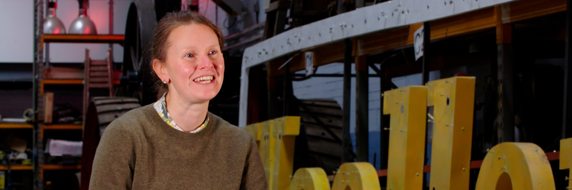 A photograph of a adult sitting in a workshop. There is a large lettered sign next to them (the letters o-l-l-e can be seen) and there are many objects behind them including industrial lights, wheels and ladders. 