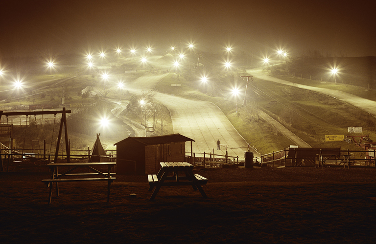 A photograph of a ski slope in Sheffield. There are lots of lightpoles, all with their lights shining brightly, creating a starburst effect in the cameras lens. A few different ski slopes are visible between the lights. A playground and picnic benches are visible in the foreground.