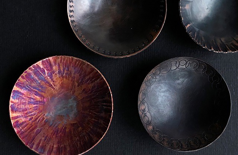 Four copper bowls. Three are dark grey with decorative details around the edges. One is orange and purple in colour.