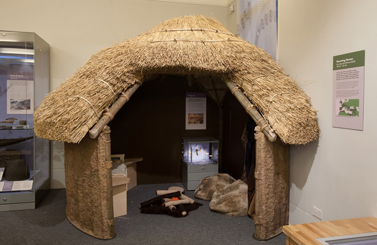 A thatched small round building with a large doorway through which you can see the interior including a replica camp fire. There are a couple of display cases inside and outside the roundhouse which hold artefacts and information sheets. 