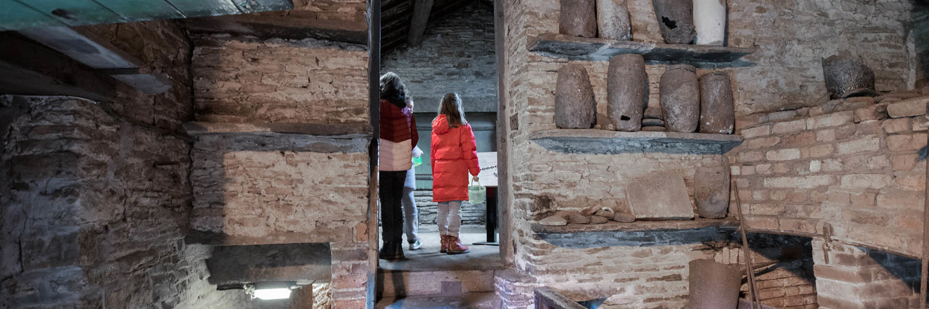 Visitors seen through a doorway with a display of old crucibles on very old wall shelves off to one side.