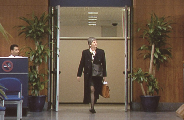 A video still of a smartly dressed person walking through a doorway marked "International Arrivals" at an airport terminal. To the left of the doorway there is a a person is sitting at a desk with a large "No Smoking Sign" on the front of it and chairs in front and to the right of the doorway a person is waiting behind a barrier.  