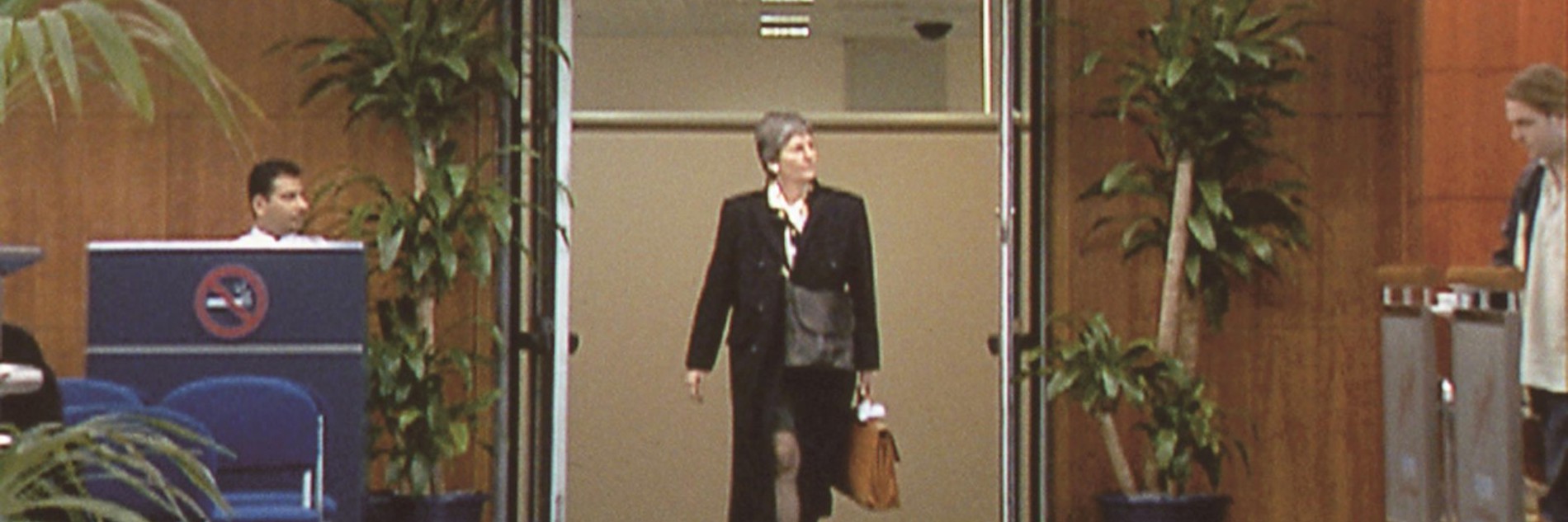 A video still of a smartly dressed person walking through a doorway marked "International Arrivals" at an airport terminal. To the left of the doorway there is a a person is sitting at a desk with a large "No Smoking Sign" on the front of it and chairs in front and to the right of the doorway a person is waiting behind a barrier.  