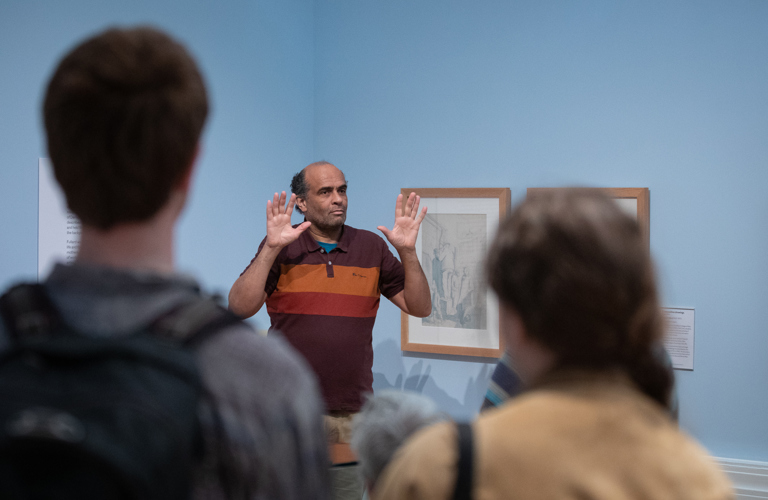 A photograph of someone delivering a gallery tour using British Sign Language. A male tour guide is shown with both hands held up, palms facing forward. He is photographed against the blue walls of the gallery.