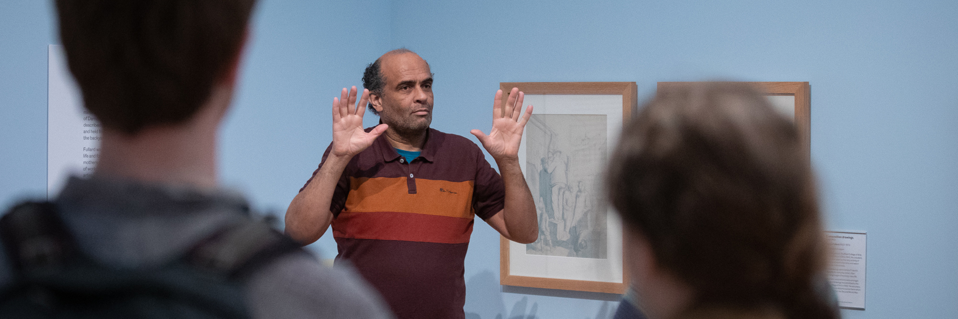 A photograph of someone delivering a gallery tour using British Sign Language. A male tour guide is shown with both hands held up, palms facing forward. He is photographed against the blue walls of the gallery.