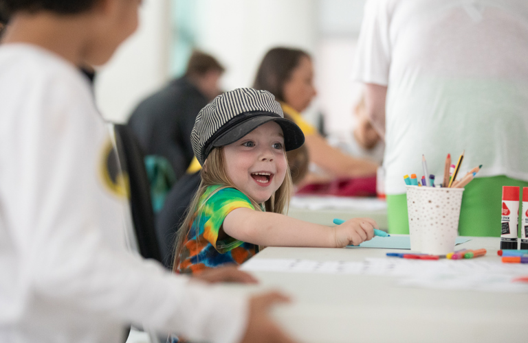 A photograph of a young girl in a stripy black and white hat smiling widely as she draws. There is a paper cup filled with pens and pencils, two gluesticks, and paper on the table in front of her.