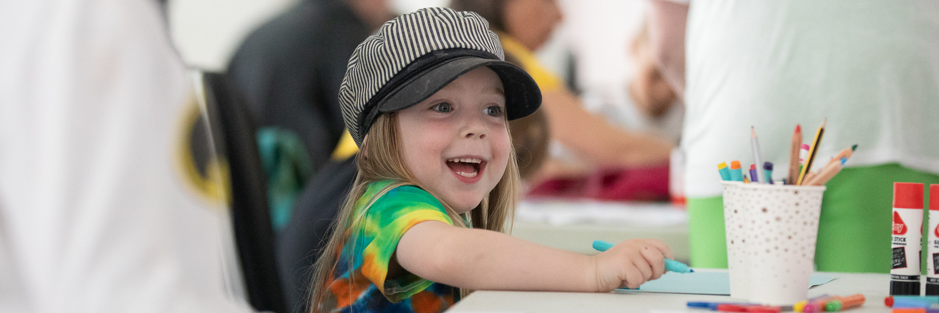 A photograph of a young girl in a stripy black and white hat smiling widely as she draws. There is a paper cup filled with pens and pencils, two gluesticks, and paper on the table in front of her.