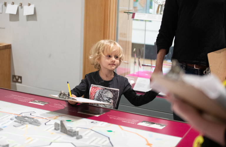A photograph of a young child with blonde curly hair looking around a museum holding a clipboard and a pencil. There is a map in front of him with 3D elements (like houses) on it.