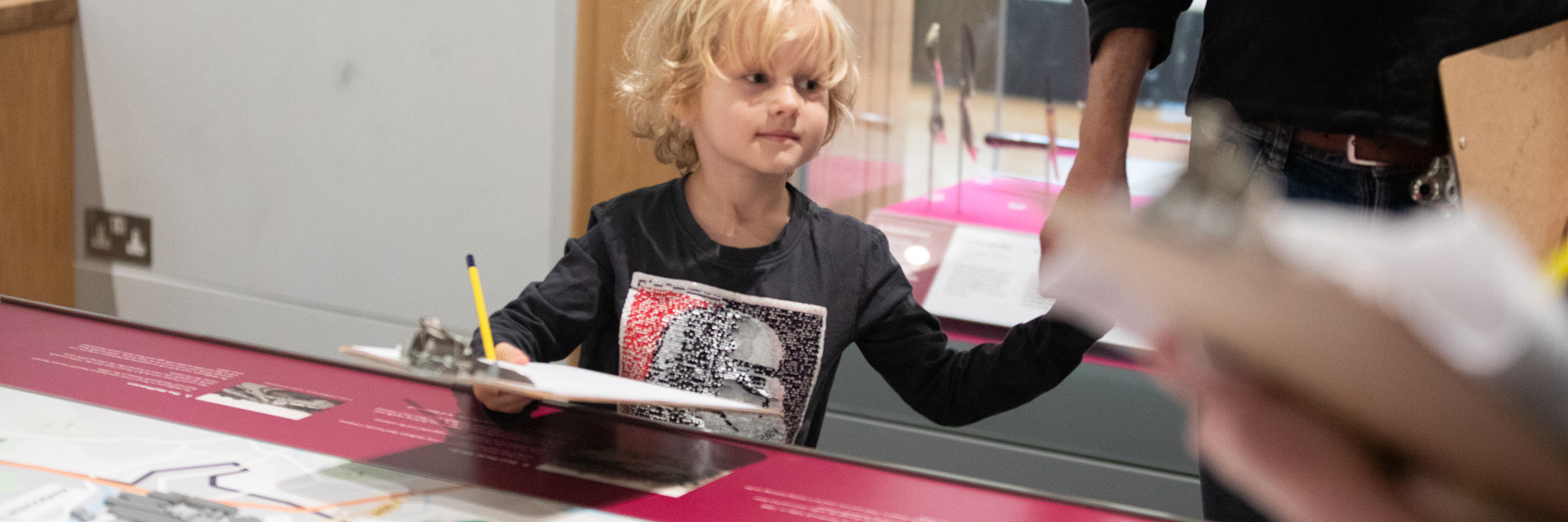 A photograph of a young child with blonde curly hair looking around a museum holding a clipboard and a pencil. There is a map in front of him with 3D elements (like houses) on it.