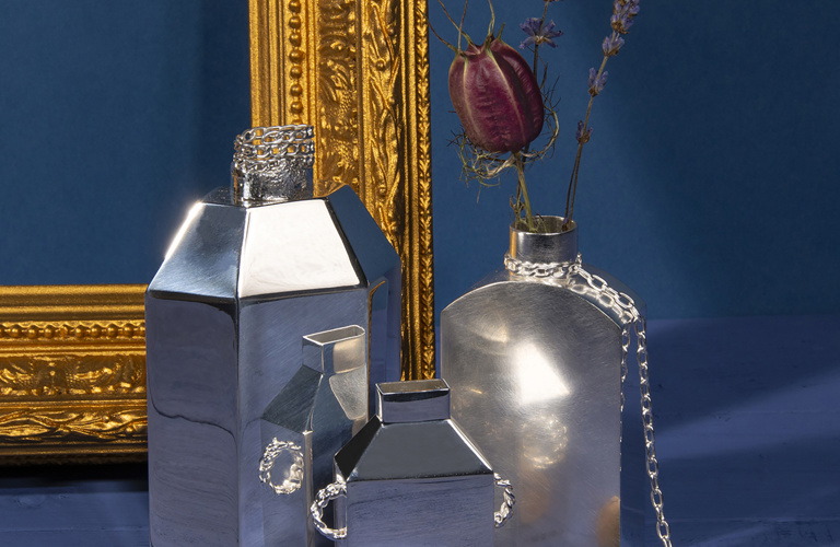 Three metal vases sitting next to a golden frame on a blue background. One of the vases is holding purple flowers. 