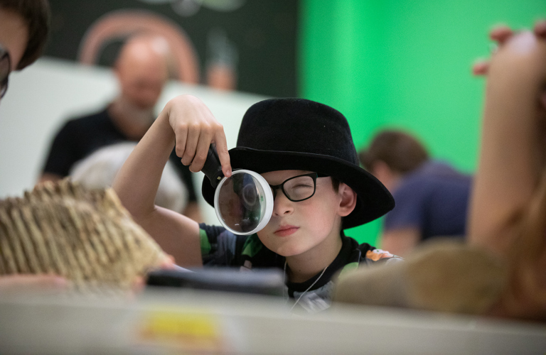 A photograph of a young boy wearing a black fedora hat, wearing black glasses and looking through a large white magnifying glass. Natural histories specimens are on the table in front of him.