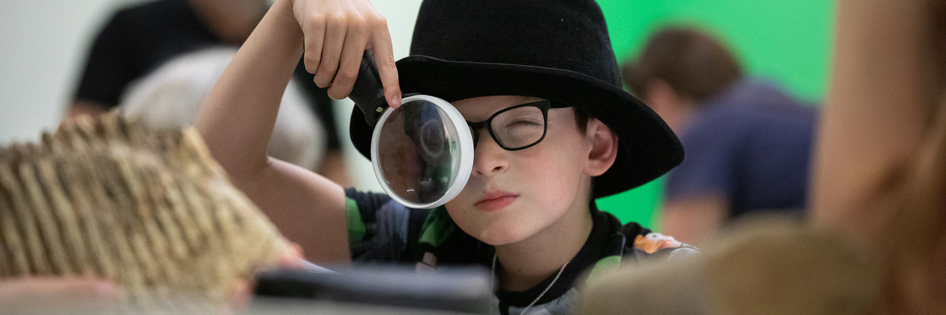 A photograph of a young boy wearing a black fedora hat, wearing black glasses and looking through a large white magnifying glass. Natural histories specimens are on the table in front of him.