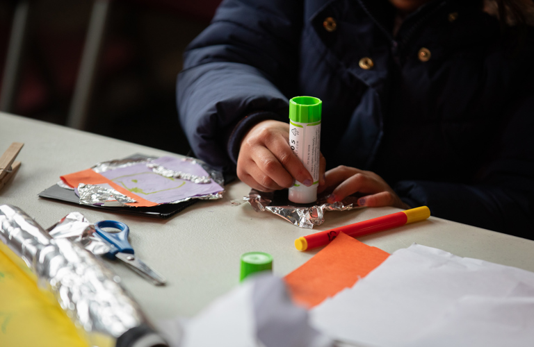 A photograph of a child's hands crafting with a glue stick, paper and aluminium foil. There is also a pair of safety scissors and a felt tip pen on the table.