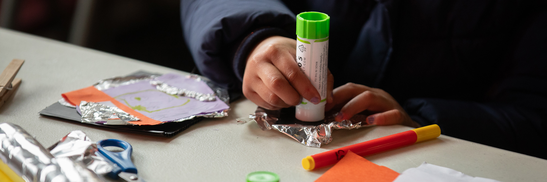 A photograph of a child's hands crafting with a glue stick, paper and aluminium foil. There is also a pair of safety scissors and a felt tip pen on the table.