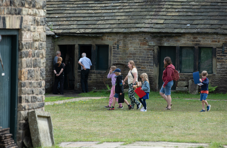 A group of adults and children walk across a grassed area in front of a row of single story historic stone buildings. 