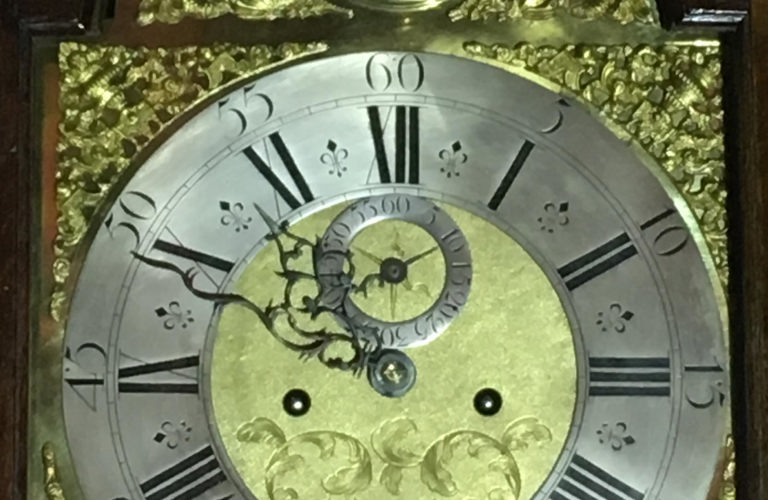 A close up of an antique clock face showing ornate hands, floral decorative motifs and Roman numerals.