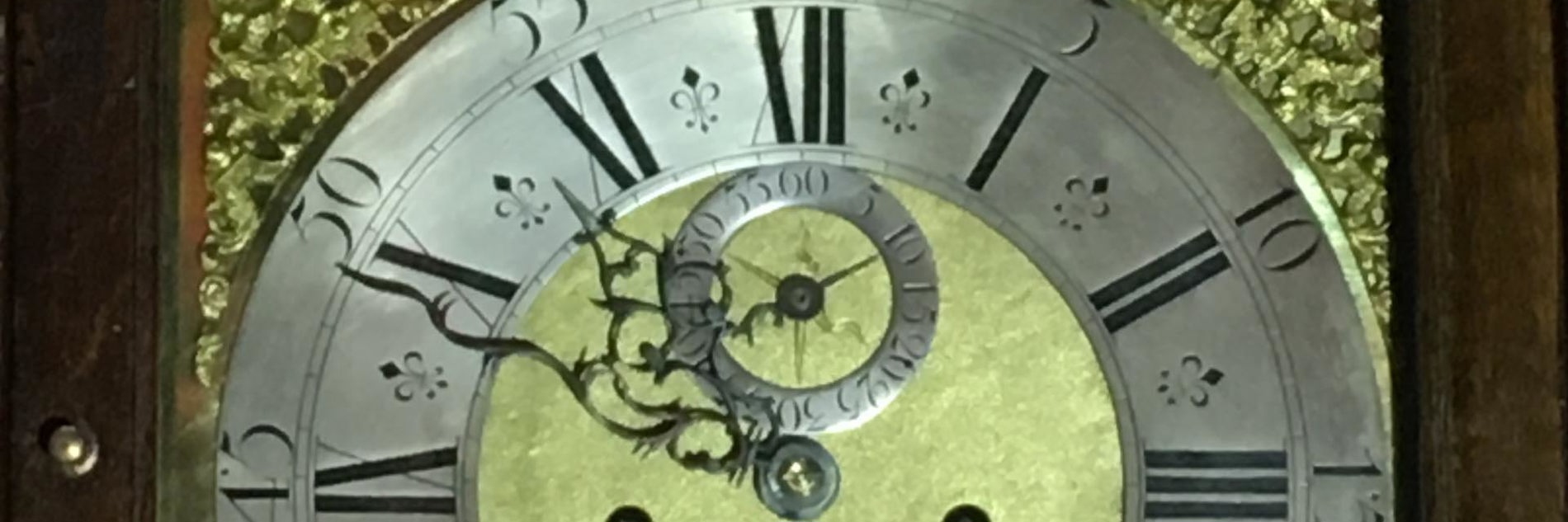 A close up of an antique clock face showing ornate hands, floral decorative motifs and Roman numerals.