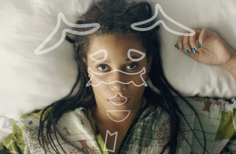 This is the alt text they want using on the image: A Black woman in her 30s lying on a bed, we see her from above and she looks up at us intensely. Her left hand with green painted nails lays next to her head on the white pillow, hair in singles on her shoulders, over a green patterned robe. She is staring not at us, but at something floating above her face, whether it is actually there or not. It is a series of symbols, formed from white outlines, in an almost-symmetrical pattern. They appear to be in motion