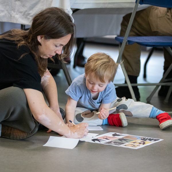 A photograph of a woman and a young boy sat on the floor with a sheet of bird images in front of them. They are both sketching on another piece of paper.