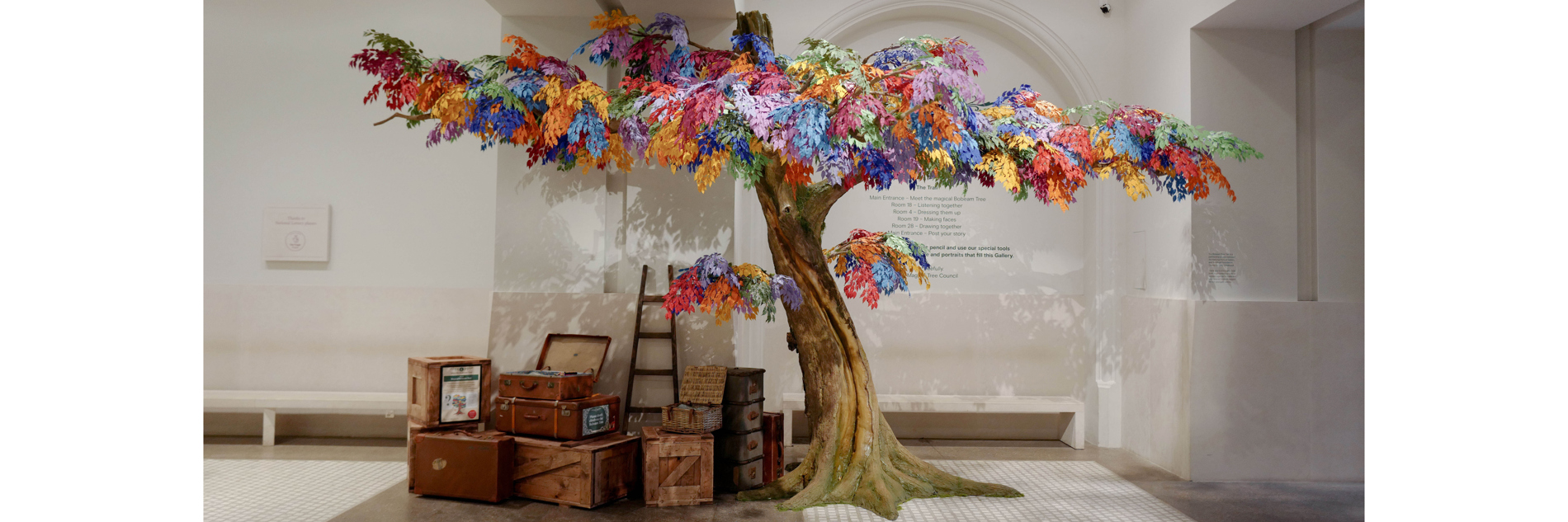 A tree with multi-coloured leaves in a gallery space next to a number of crates and boxes 