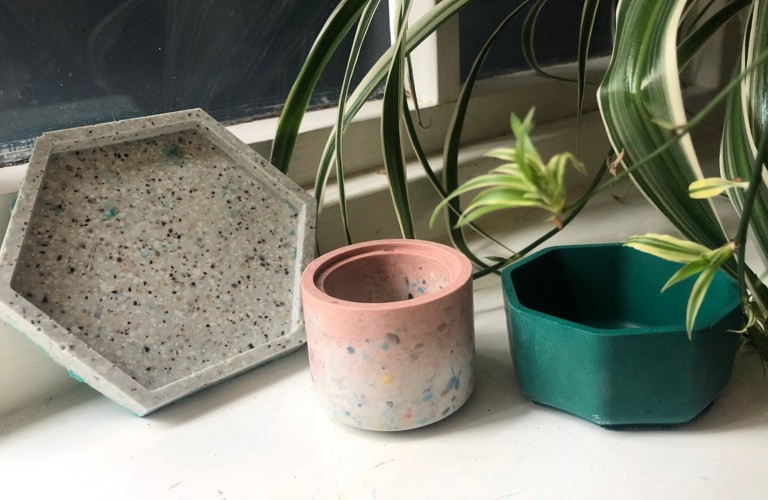 Three plant pots lined up on a windowsill with a houseplant partially visible in the top left corner. From left to right, a grey hexagonal plant pot saucer, a pink and white cylindrical plant pot and a green hexagonal plant pot. 