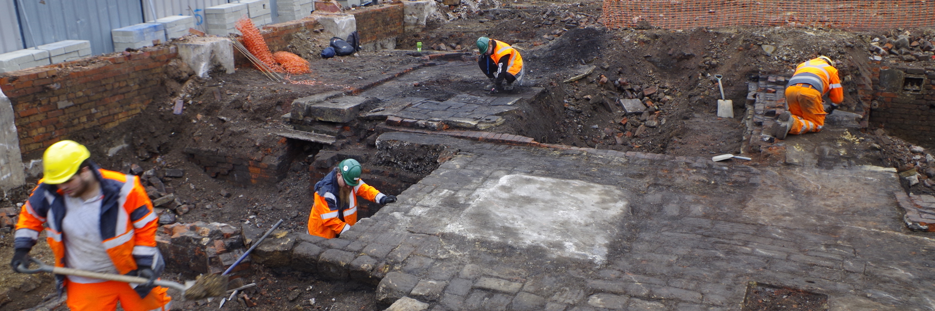 A photograph of four workers in orange high vis and hard hats excavating the Sheffield Castle dig site. There are boards and scaffolding surrounding the site and various stone work paths that the workers are carefully working around.