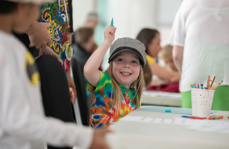 A photograph of a smiling child sitting at a table and holding a pen. On the table there is a paper, pens and a pot of pens.  Other children and adults can be seen in the background. 