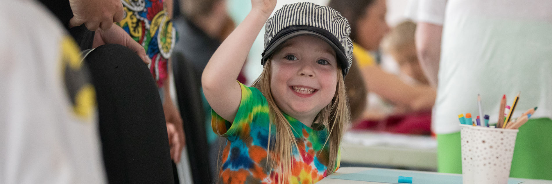 A photograph of a smiling child sitting at a table and holding a pen. On the table there is a paper, pens and a pot of pens.  Other children and adults can be seen in the background. 