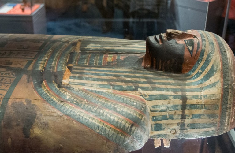 A photograph of the mummy in Weston Park Museum. The sarcophagus is decorated with blue and orange lines, with bold black lines around the eyes and mouth.