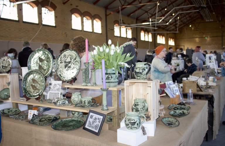 A photograph of a row of tables displaying a selection of ceramics on shelves and display units. Several adults are stood behind the tables. 