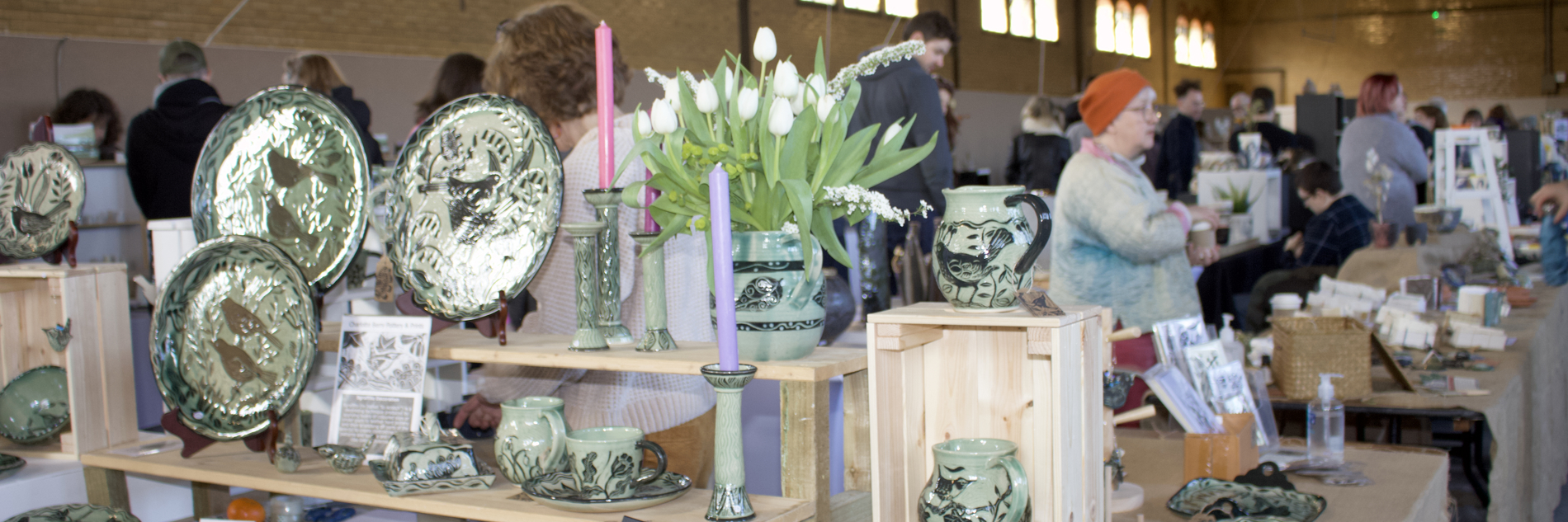 A photograph of a row of tables displaying a selection of ceramics on shelves and display units. Several adults are stood behind the tables. 