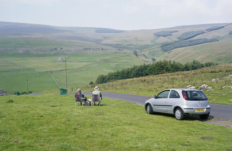  A photograph showing a rural landscape. In the foreground two people sit in folding chairs next to a road and a parked car.  