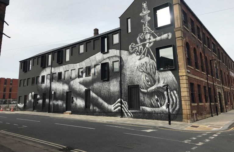 A photograph of a large building with a mural painted on the side. The background is matte black, and humanoid figure is shown lying down, painted in greyscale.
