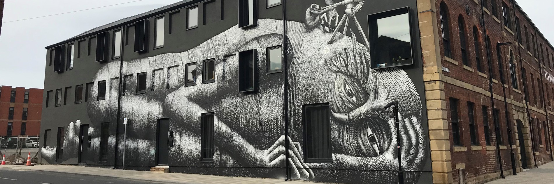 A photograph of a large building with a mural painted on the side. The background is matte black, and humanoid figure is shown lying down, painted in greyscale.