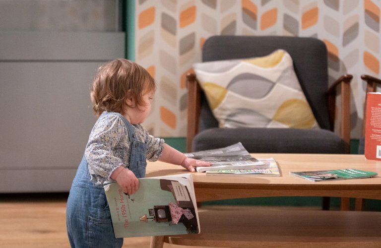 A photograph of a toddler arranging magazines on a low wooden table in Weston Park Museum.