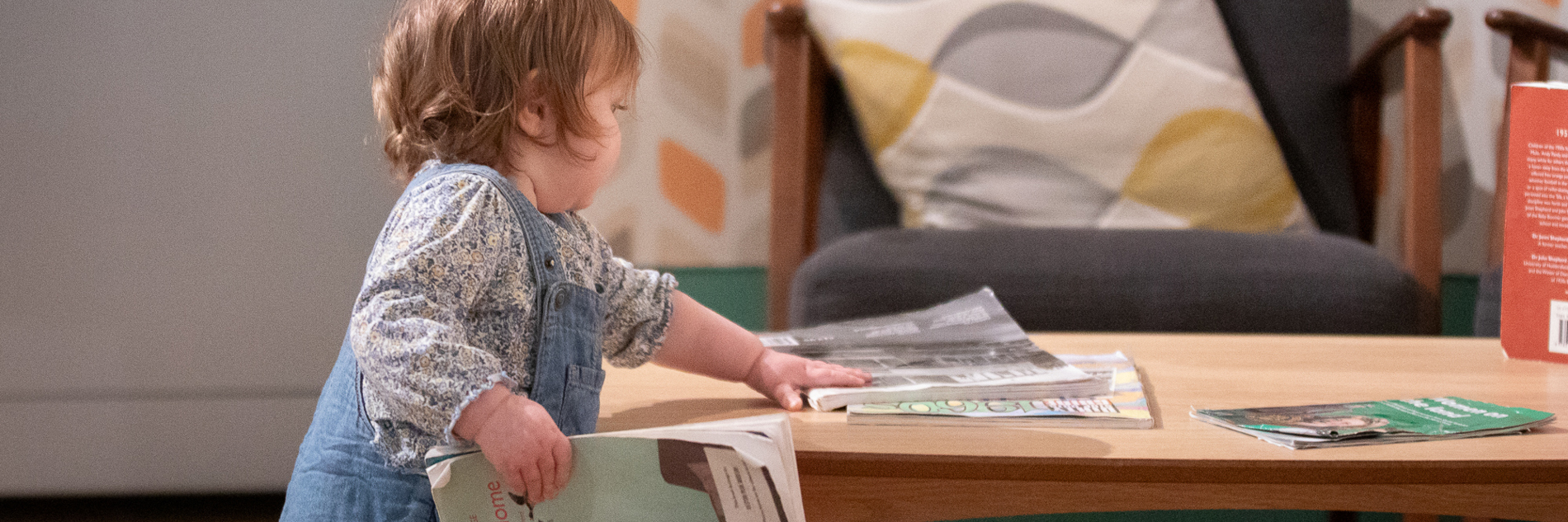 A photograph of a toddler arranging magazines on a low wooden table in Weston Park Museum.