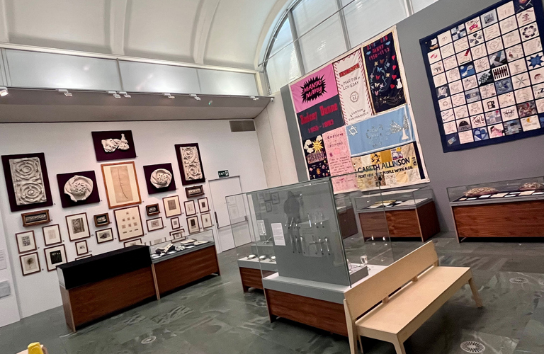 A modern gallery space with a curved ceiling> on the right hand wall are two embroidered patchwork quilts. On the left hand way are a series of works on paper and plaster reliefs. In the foreground are a series of display cases and a modern light wood bench. 