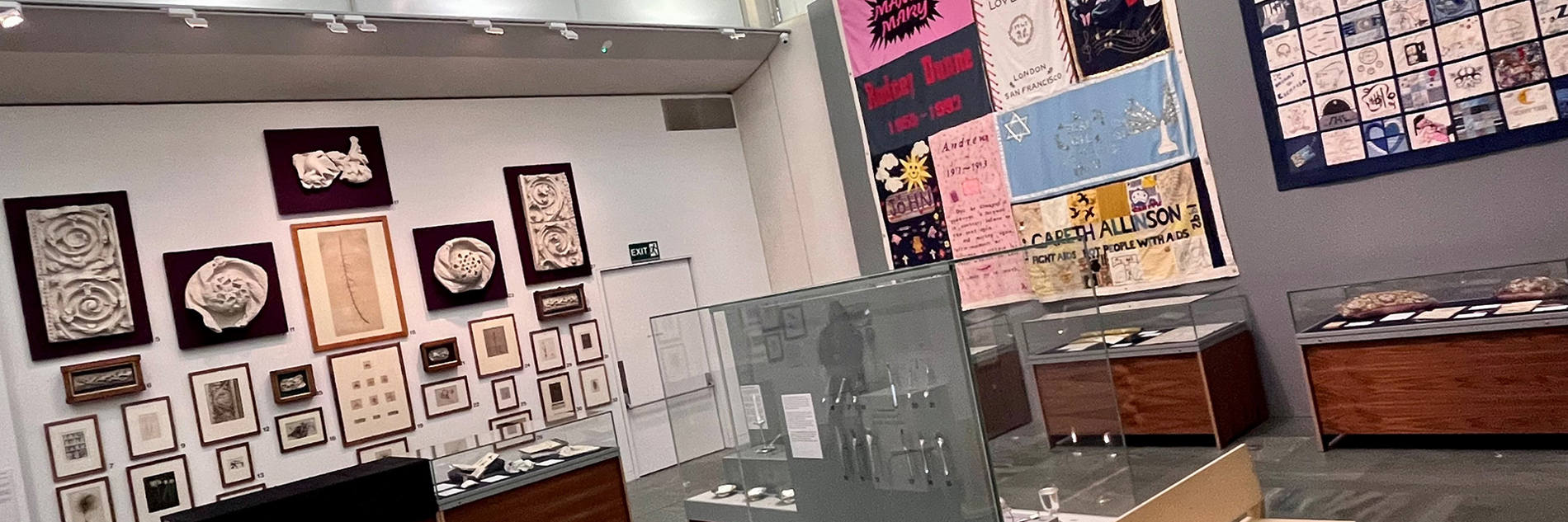 A modern gallery space with a curved ceiling> on the right hand wall are two embroidered patchwork quilts. On the left hand way are a series of works on paper and plaster reliefs. In the foreground are a series of display cases and a modern light wood bench. 