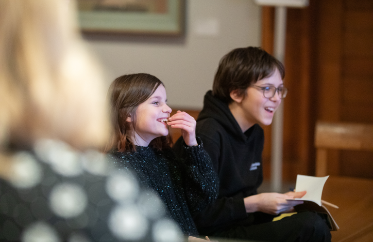 A photograph of two smiling children sitting on a wooden bench in an art gallery. 