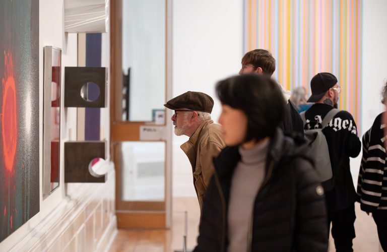 A photograph of two people looking at art in a gallery. One is stood upright, the other is leaning towards the wall.